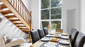 Un comedor con una mesa de madera dispuesta para cuatro, una escalera y un ventanal con vistas a árboles.