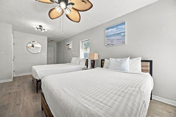 A hotel room with two beds, a ceiling fan, a wall-mounted mirror, and a beach-themed painting.