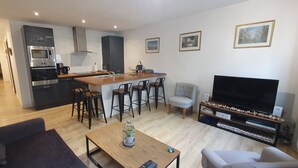 En moderne lejlighed med et køkken, spiseområde og opholdsstue. Der er et stort fjernsyn, en sofa og et sofabord.