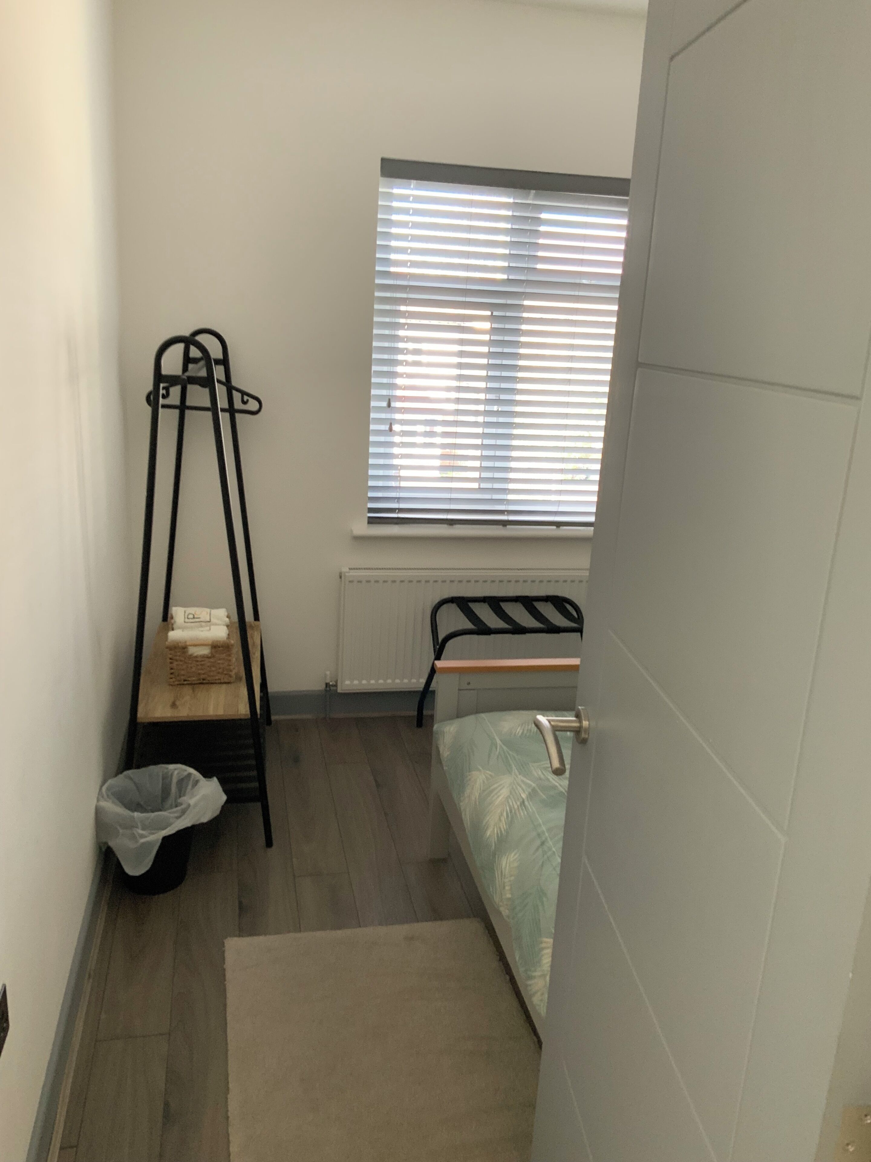 Una habitación pequeña y poco amueblada con una cama, un radiador, una ventana con persianas, una cubierta para radiador, un estante pequeño y un cubo de basura.
