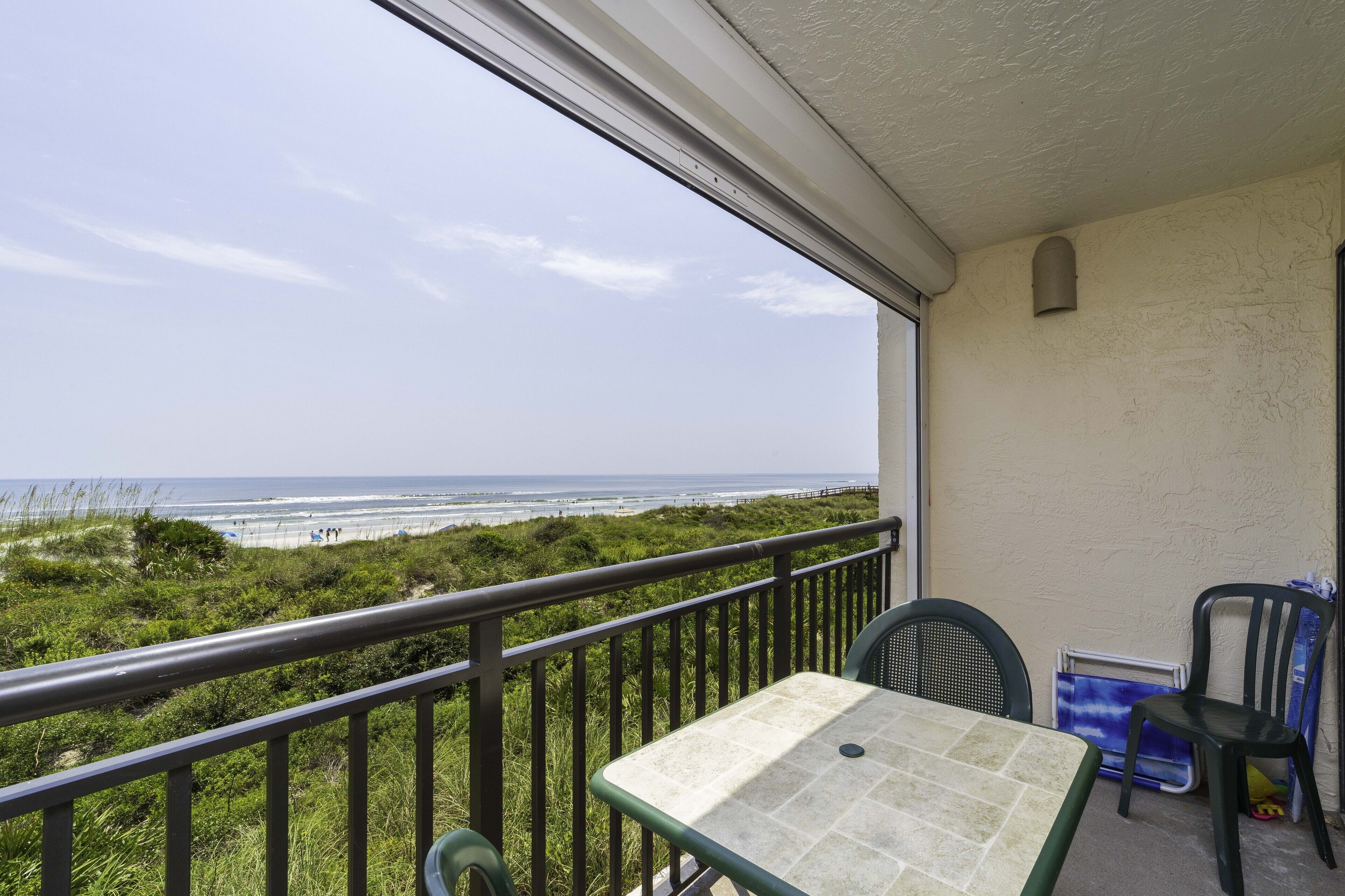 A balcony with a table and chairs, overlooking a beach with people in the distance.