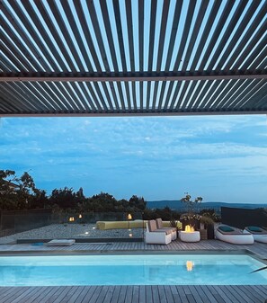 A modern outdoor pool area with a wooden deck, seating, and a pergola overhead.