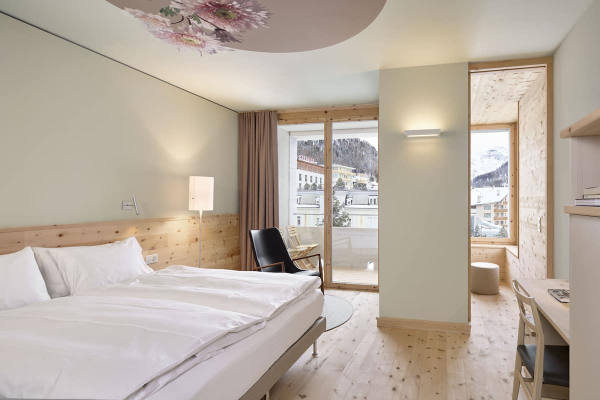 Une chambre d’hôtel avec un lit, une chaise, un bureau et une vue sur une montagne enneigée et des bâtiments.