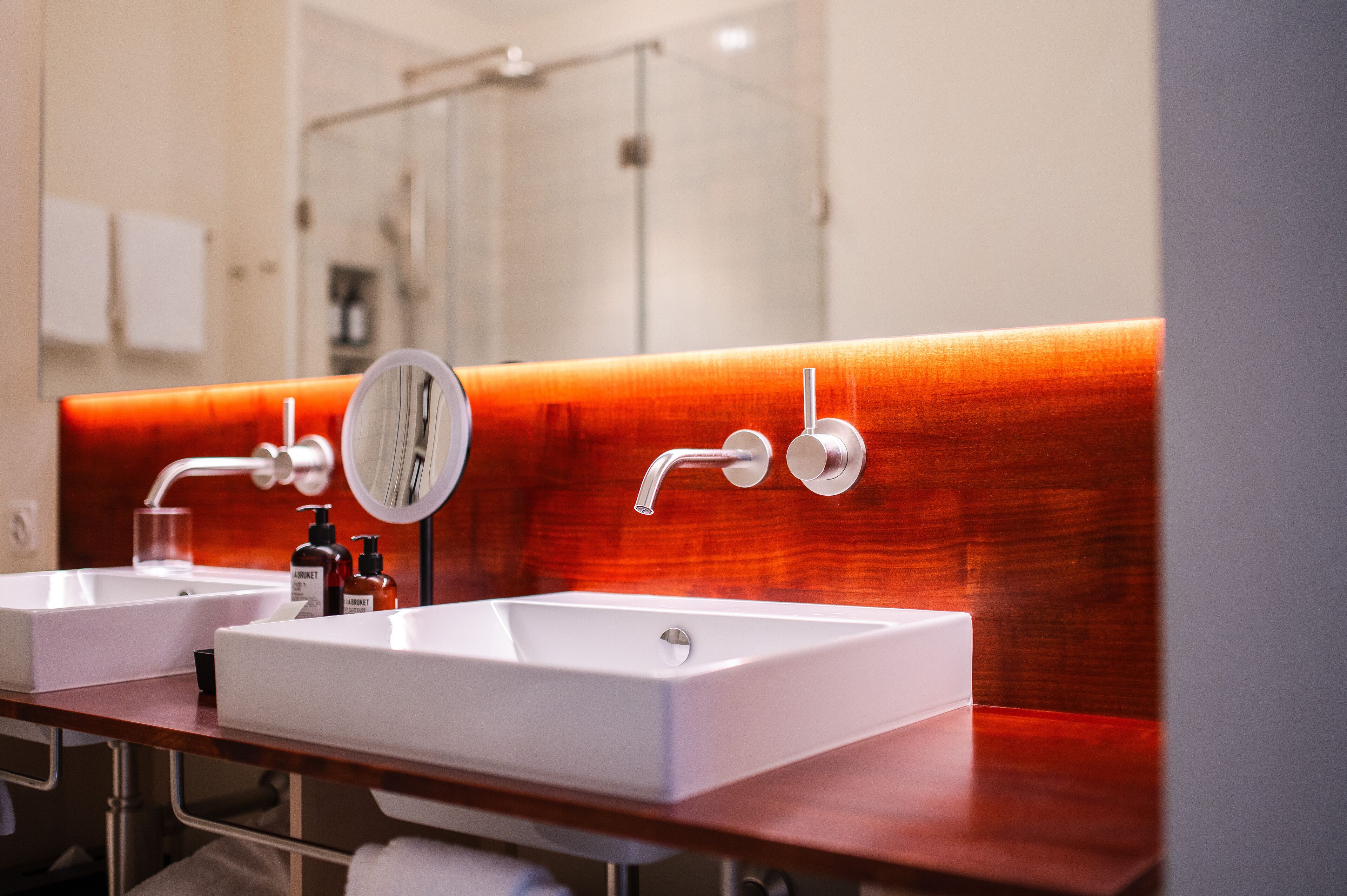 Un baño moderno con dos lavamanos blancos, un espejo y una pared de acento roja.