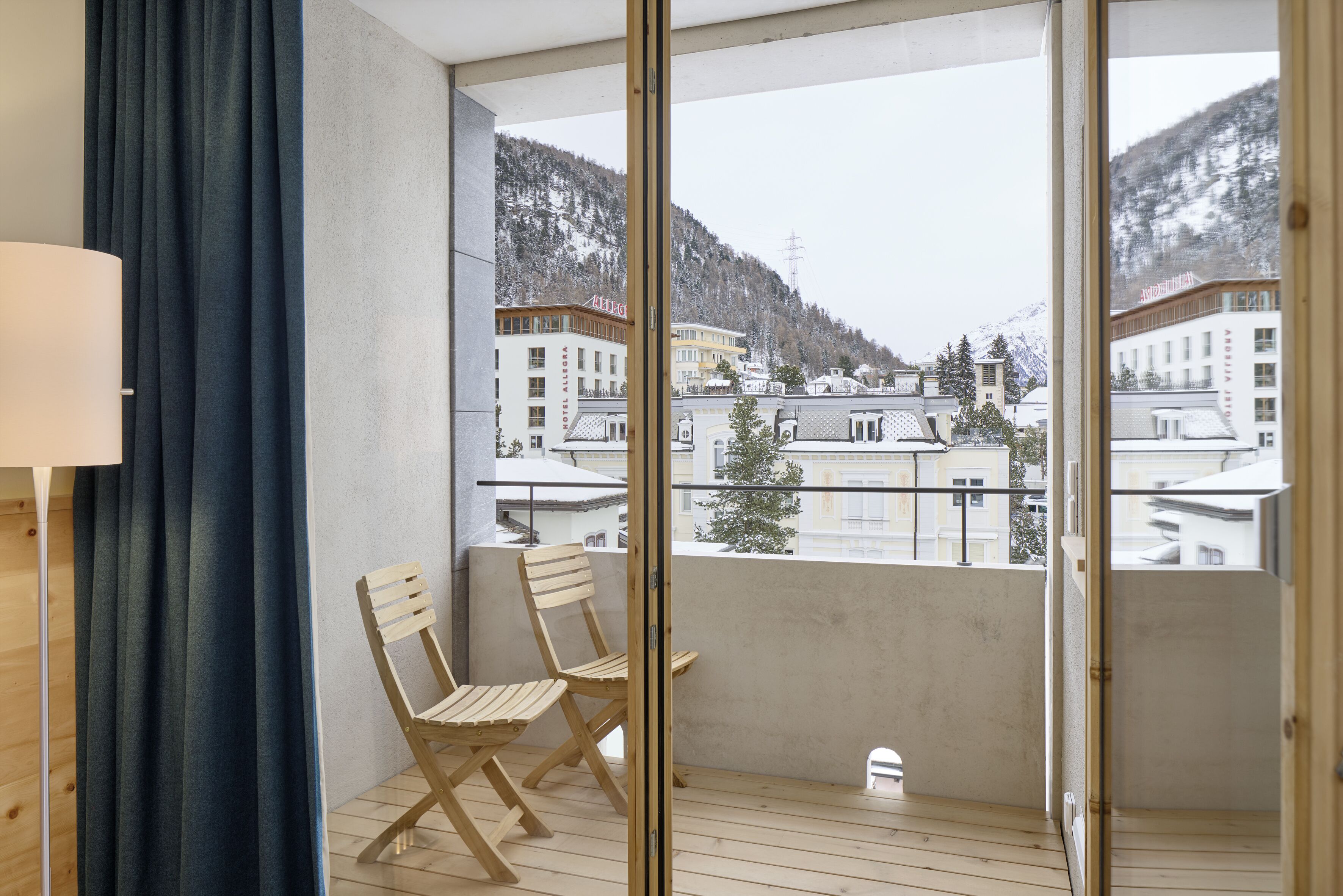 A balcony with wooden chairs, a view of buildings and mountains, and a lamp on the left.