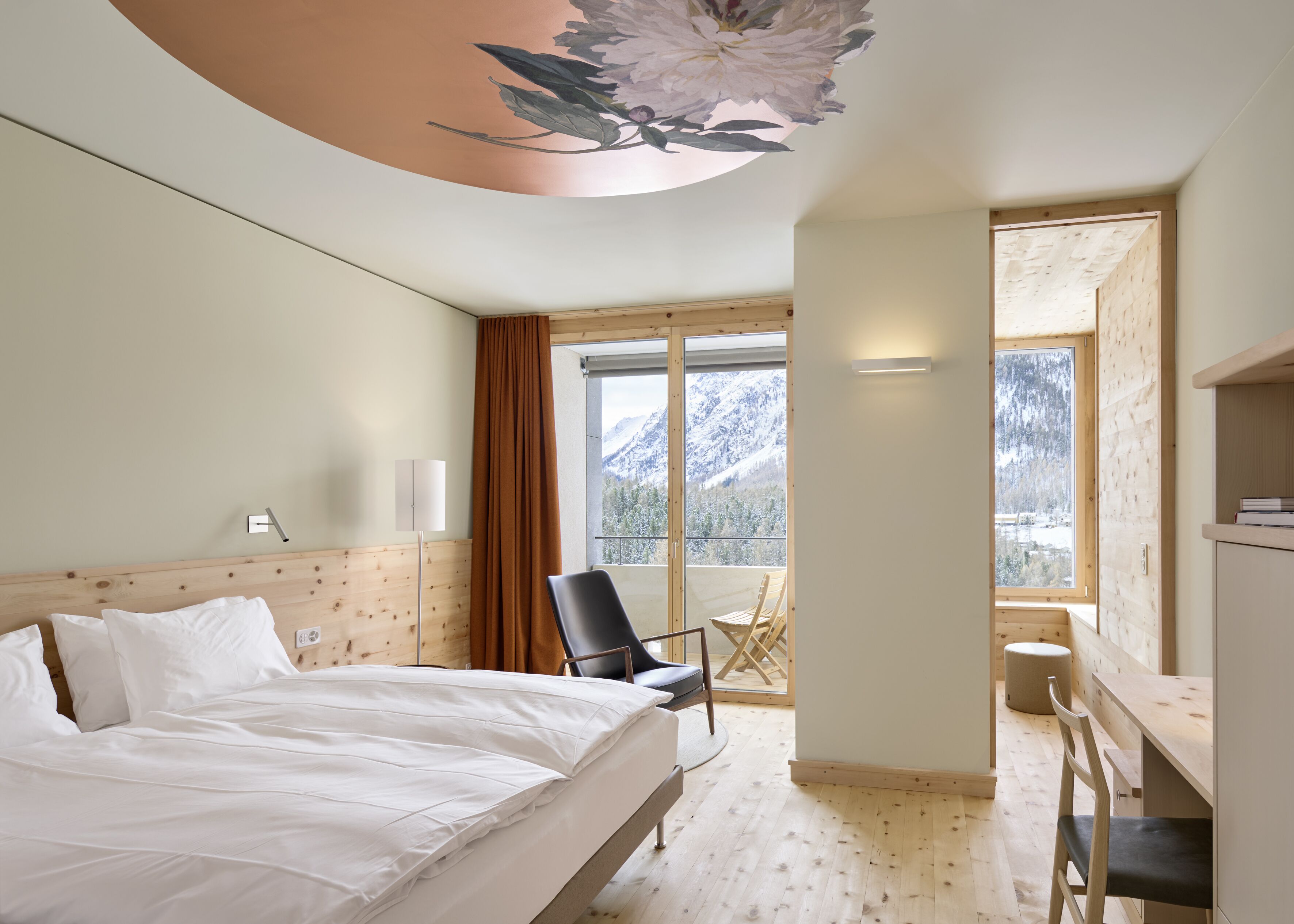 Habitación de hotel con cama, escritorio, silla y un gran ventanal con vistas a la montaña.