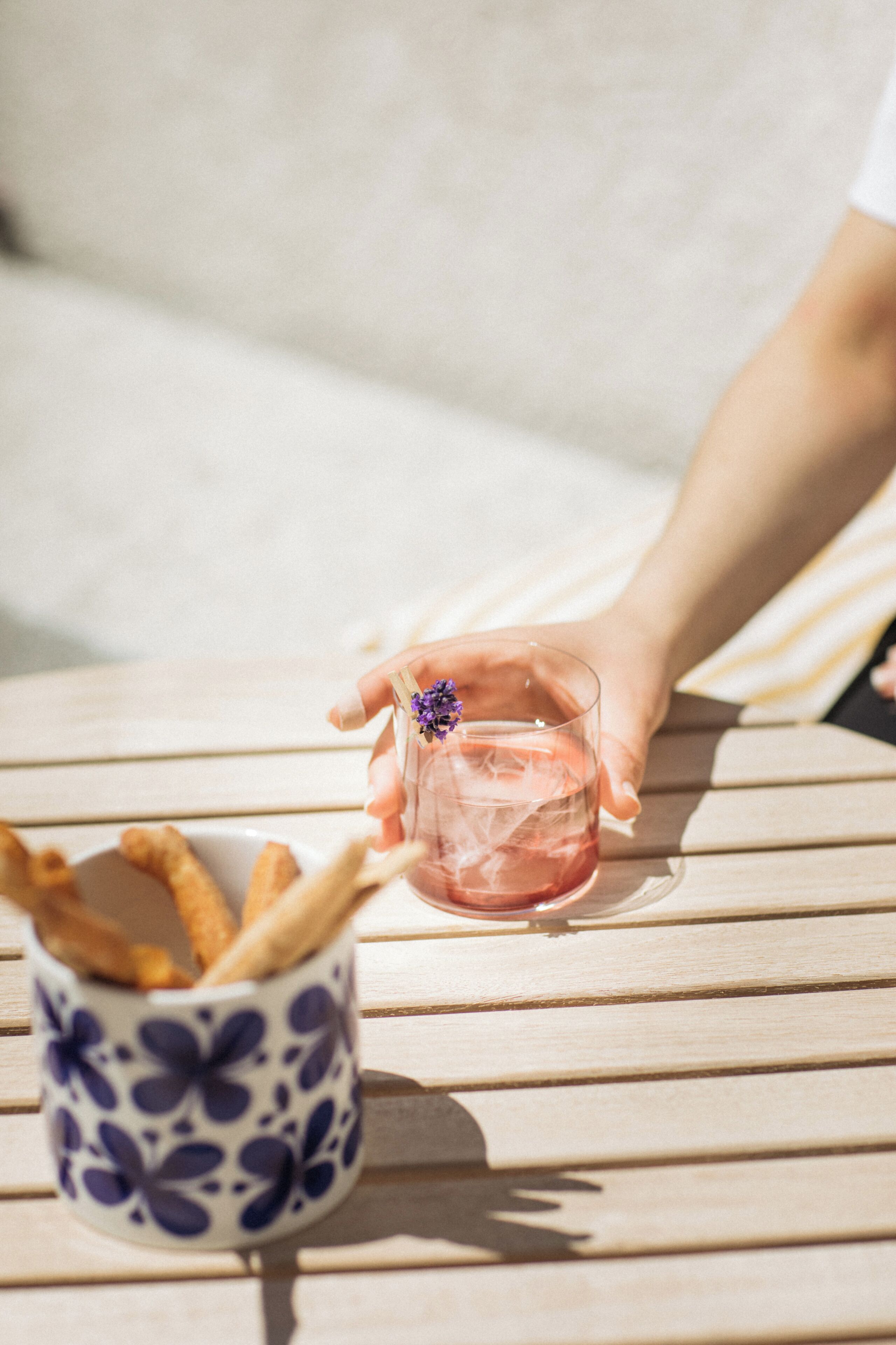 A hand holding a glass with a flower, a cup with breadsticks, and a wooden table.