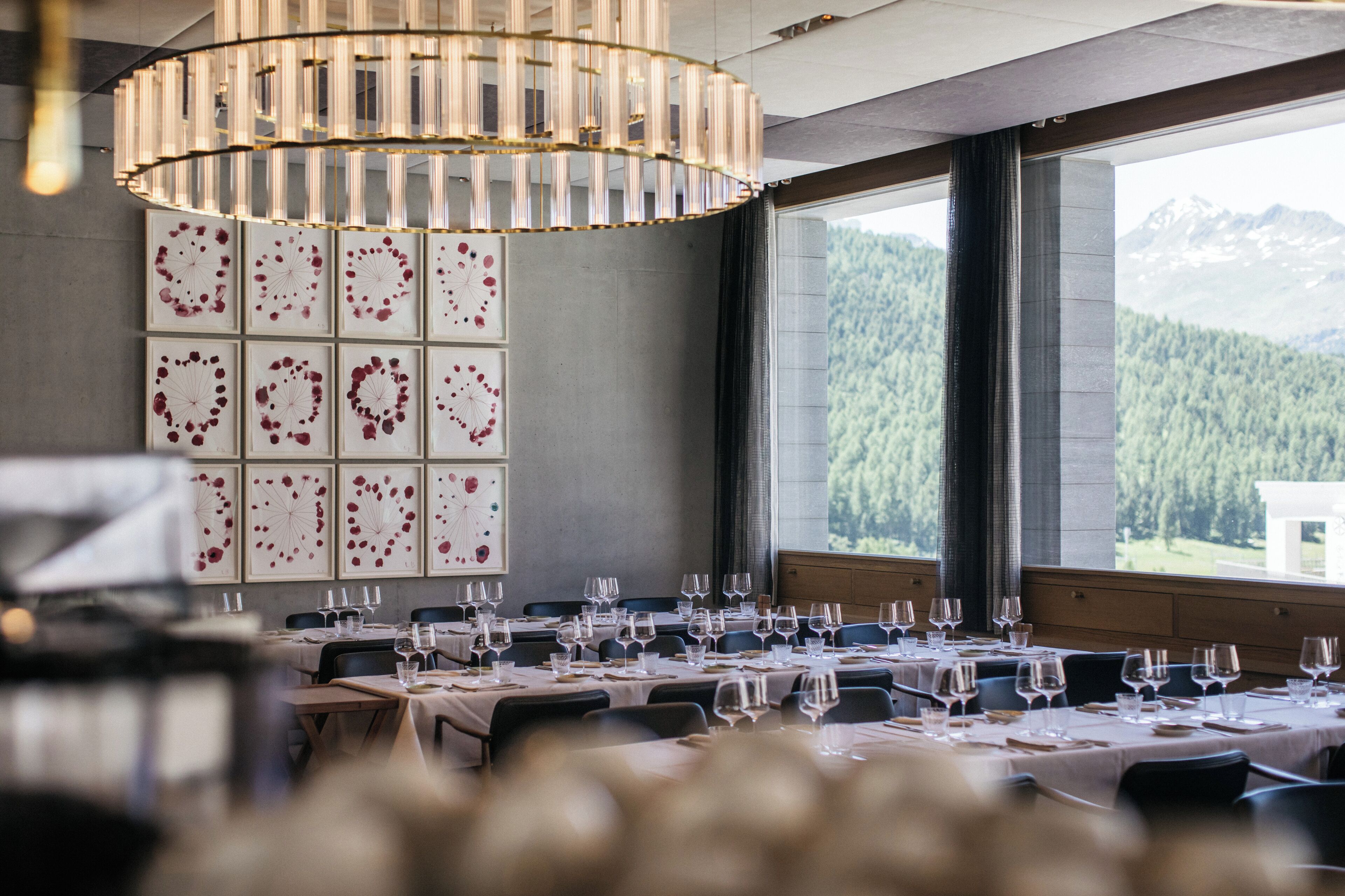 Uma sala de jantar com mesas postas para uma refeição, um grande candelabro e vista para uma cadeia montanhosa arborizada através de amplas janelas.