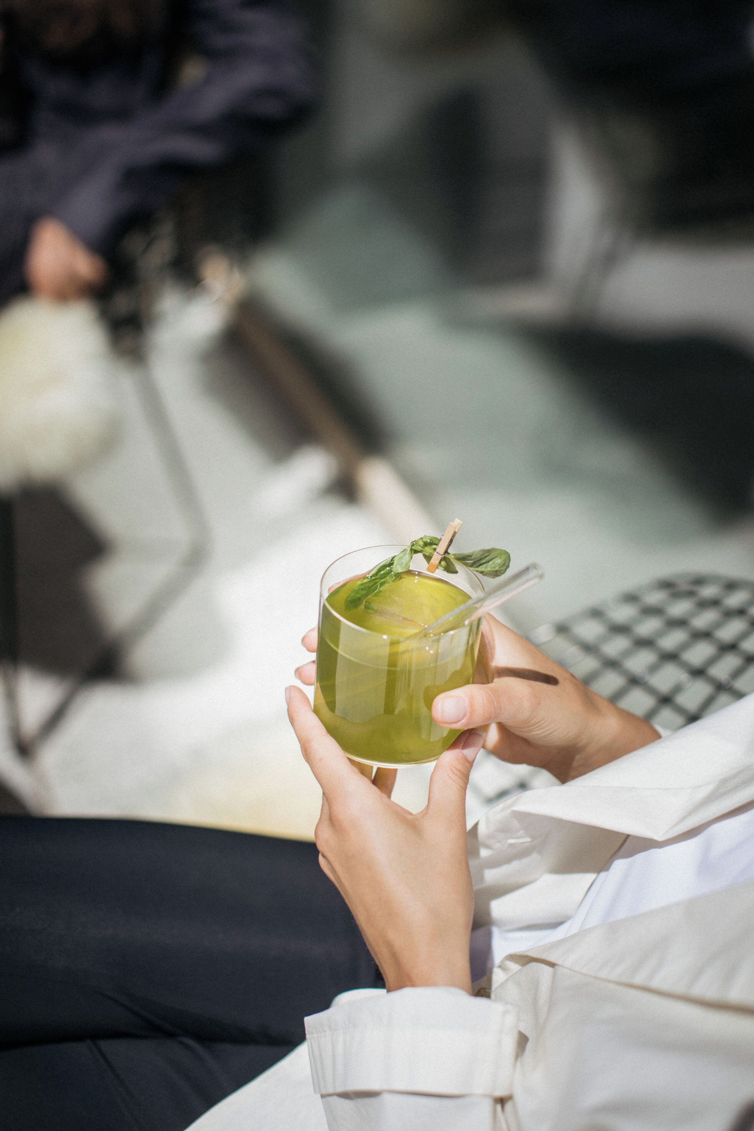 A person holding a glass with a green beverage, seated on a couch with a checkered pillow.
