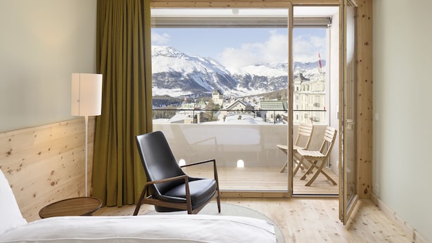 Ein Hotelzimmer mit Bett, Sessel, Lampe und Blick auf Berge sowie eine Stadt durch eine Schiebetür.