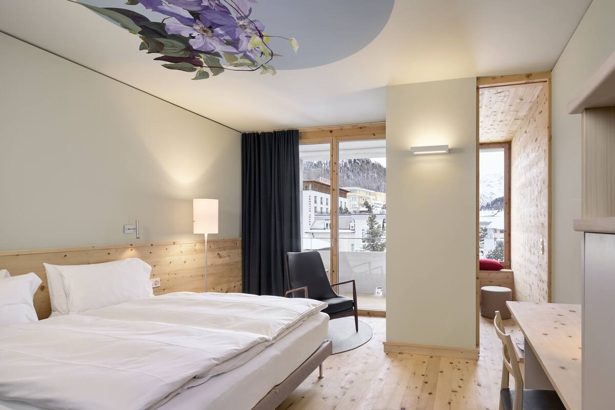 Une chambre d’hôtel avec un lit, un bureau, une chaise, une lampe et une vue sur une montagne enneigée et des bâtiments.
