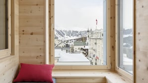 Ein Zimmer mit Holzboden, einer Bank, einem runden Hocker und einem großen Fenster mit Blick auf einen verschneiten Berg und Gebäude.