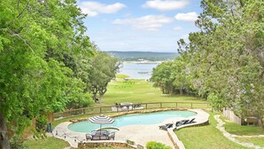 A swimming pool with a curved design, surrounded by a stone deck and a wooden fence. There is a patio area with a table and chairs, and a view of a body of water and distant land. - Above Escape LT I Lake Travis I Pool&Spa I Services I Pets (Austin)