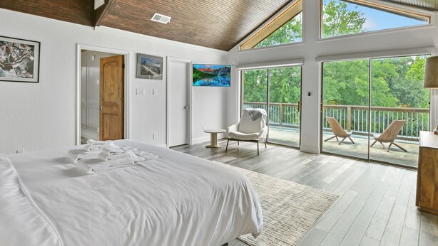 A spacious bedroom with a large bed, a chair, and a balcony with a view of trees. - Above Escape LT I Lake Travis I Pool&Spa I Services I Pets (Austin)