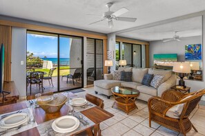 A spacious living area with a sliding glass door leading to a balcony with a view of the ocean. There are wicker chairs, a sofa, and a dining table set with plates and glasses.