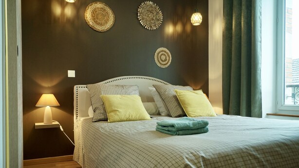 Een netjes opgemaakt bed met een geruit sprei, twee kussens en een opgevouwen handdoek. Er staat een bedlampje op een nachtkastje.