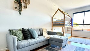 A modern living room with a grey sectional sofa, a wooden bunk bed, and a large window providing natural light. - Stunning Family Retreat Perfect Location (melbourne)