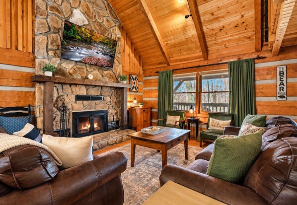 A cozy cabin interior with a stone fireplace, leather sofas, and a wooden ceiling.
