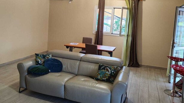 Une pièce comprenant un canapé, une table et une chaise. On y trouve une fenêtre avec des rideaux, un plafond avec des poutres en bois et un climatiseur fixé au mur.