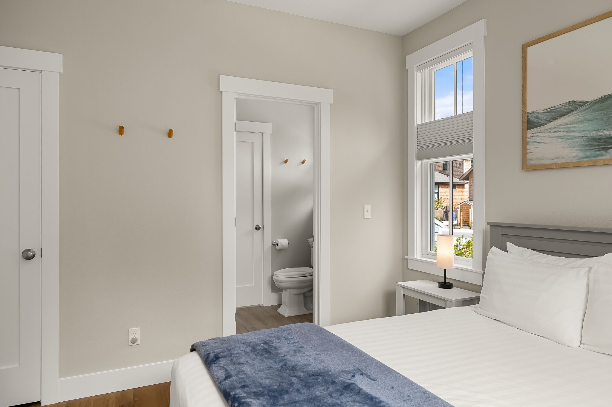 A bedroom with a bed, a bedside table with a lamp, a window with a view of the outside, and a door leading to a bathroom.