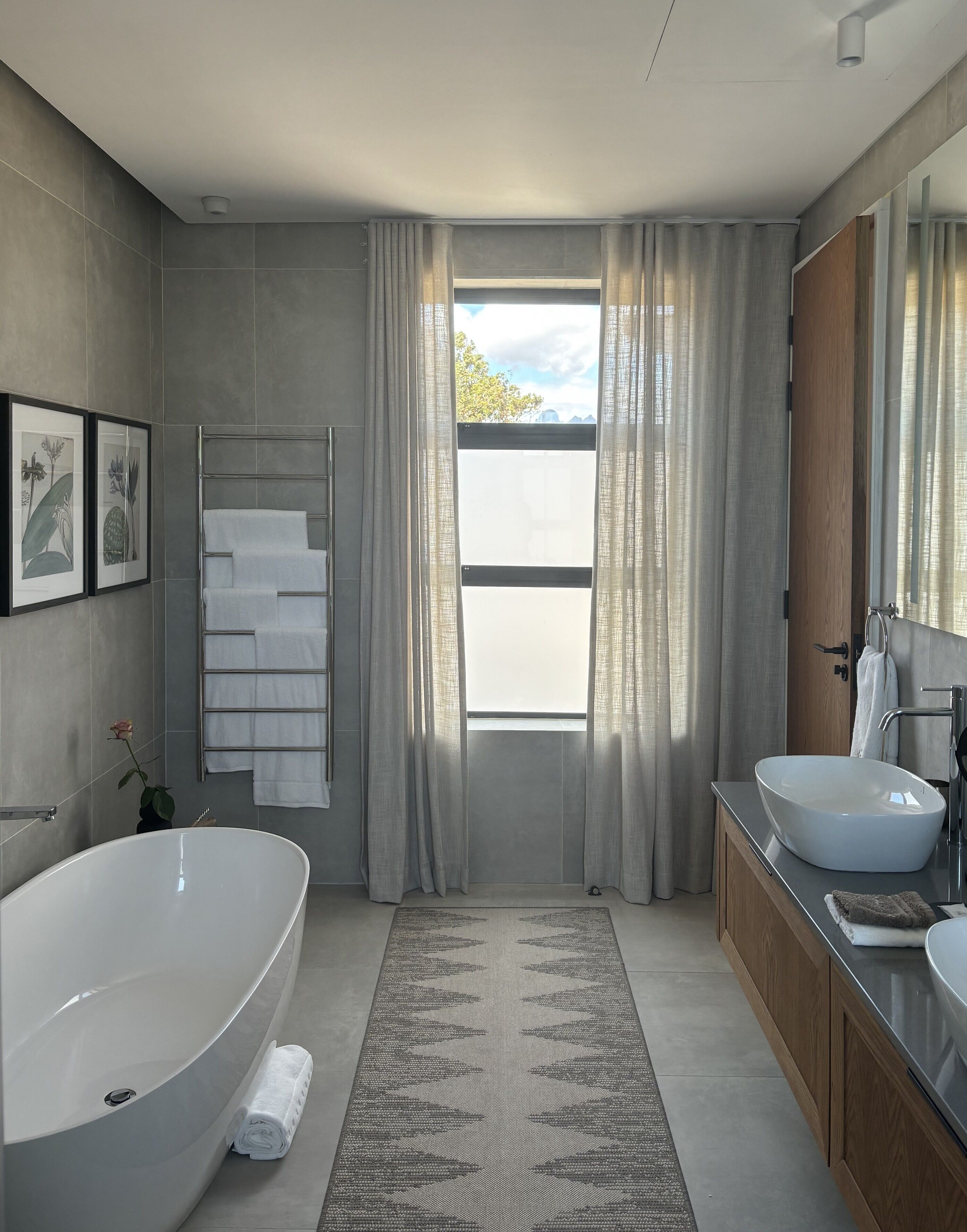 A modern bathroom with a freestanding bathtub, a vanity with a large sink, and a view of a cityscape through a window with sheer curtains.