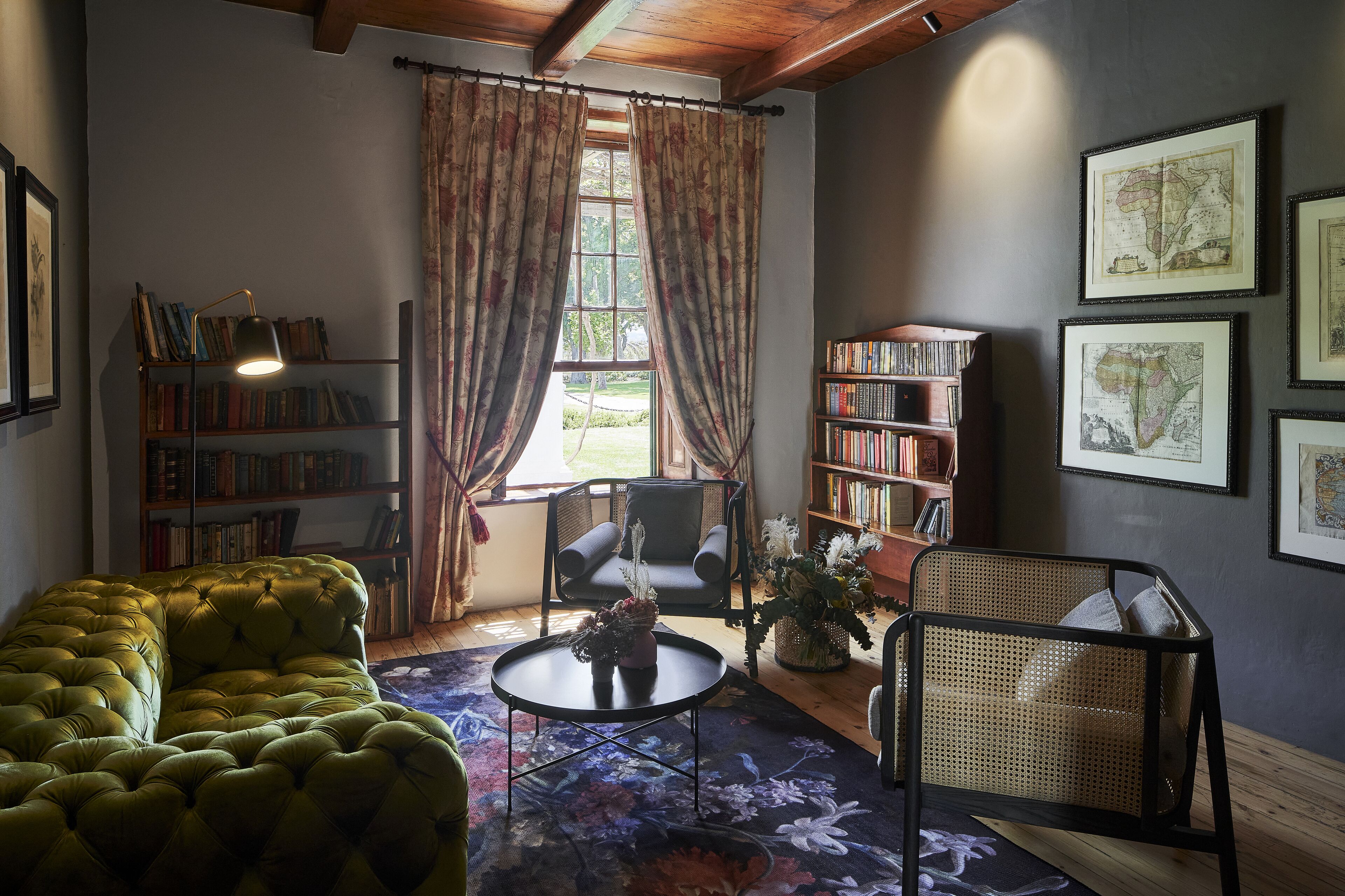 A cozy reading nook with a green sofa, a round coffee table, and a wicker chair. There are bookshelves filled with books and framed maps on the walls.