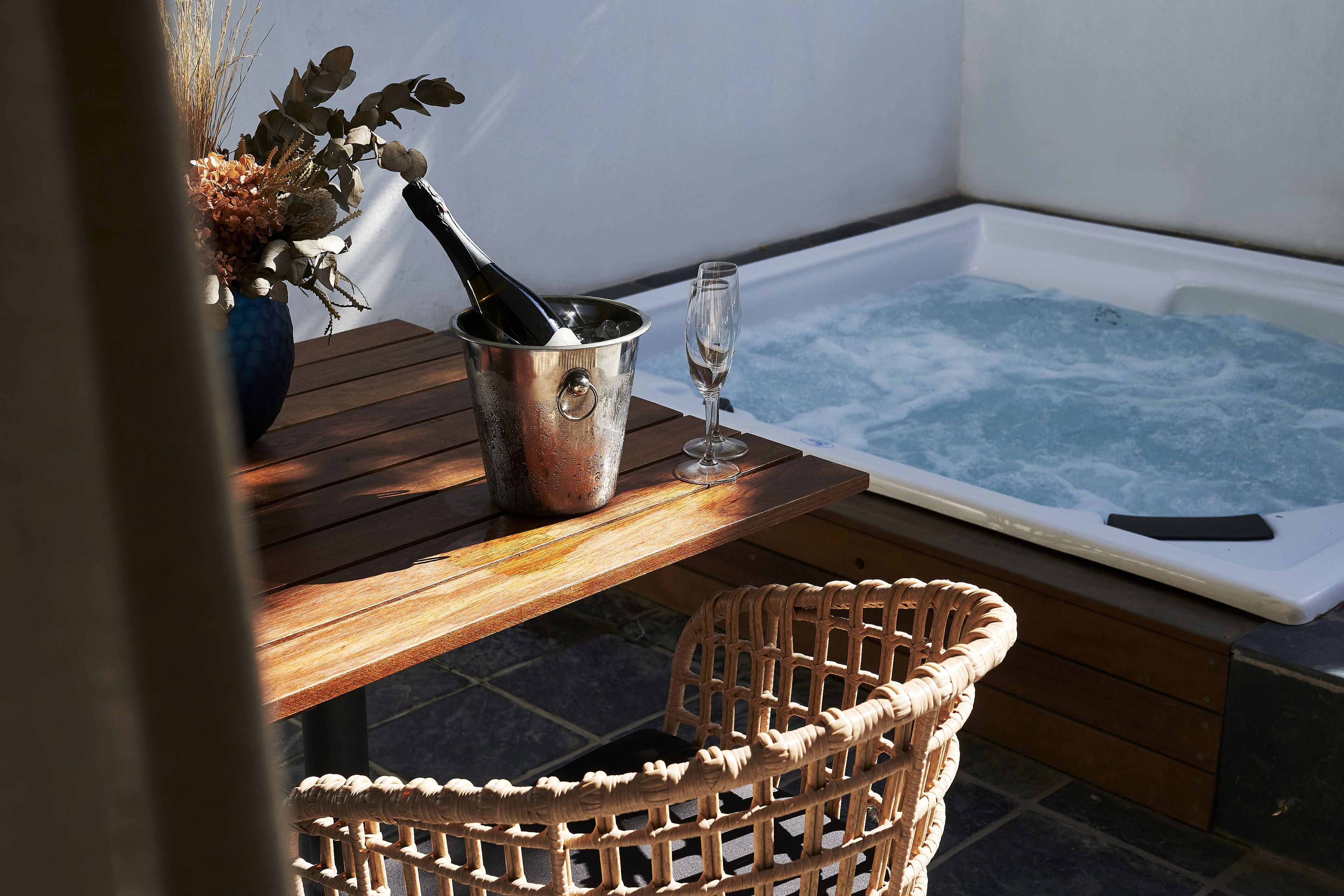 A wooden table with a metal bucket containing a bottle and a glass, next to a wicker chair, and a hot tub with a view of the sky.