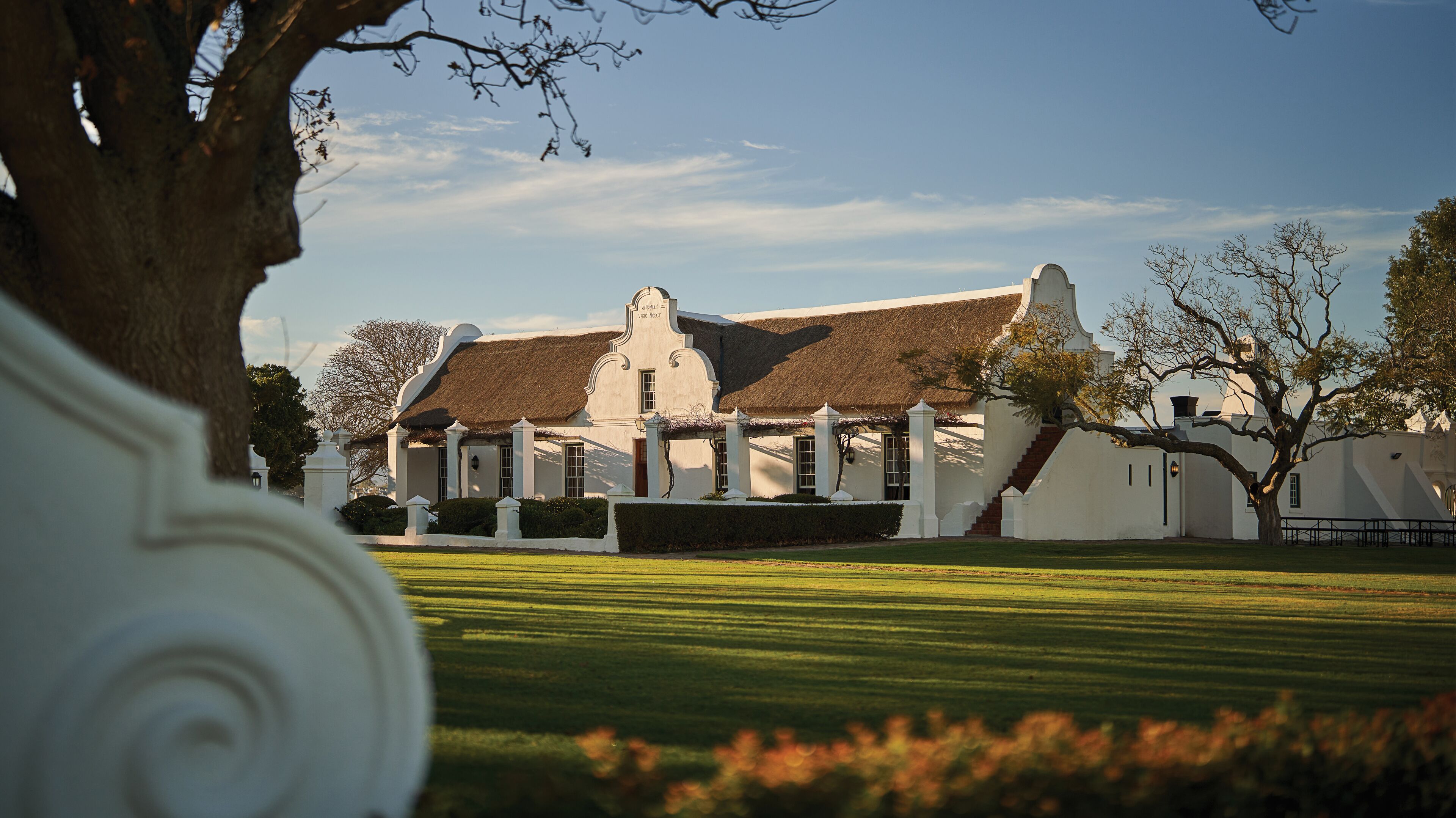 A colonial-style building with a thatched roof, surrounded by a well-maintained lawn and a large tree.