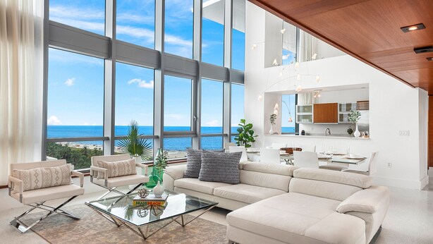 A modern living room with large windows, a glass coffee table, and a view of the ocean.