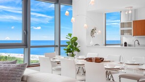 A modern dining area with a view of the sea, white furniture, and pendant lighting.