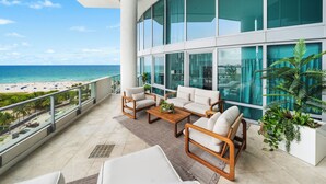A balcony with a view of the beach, furnished with a sofa, chairs, and a coffee table.
