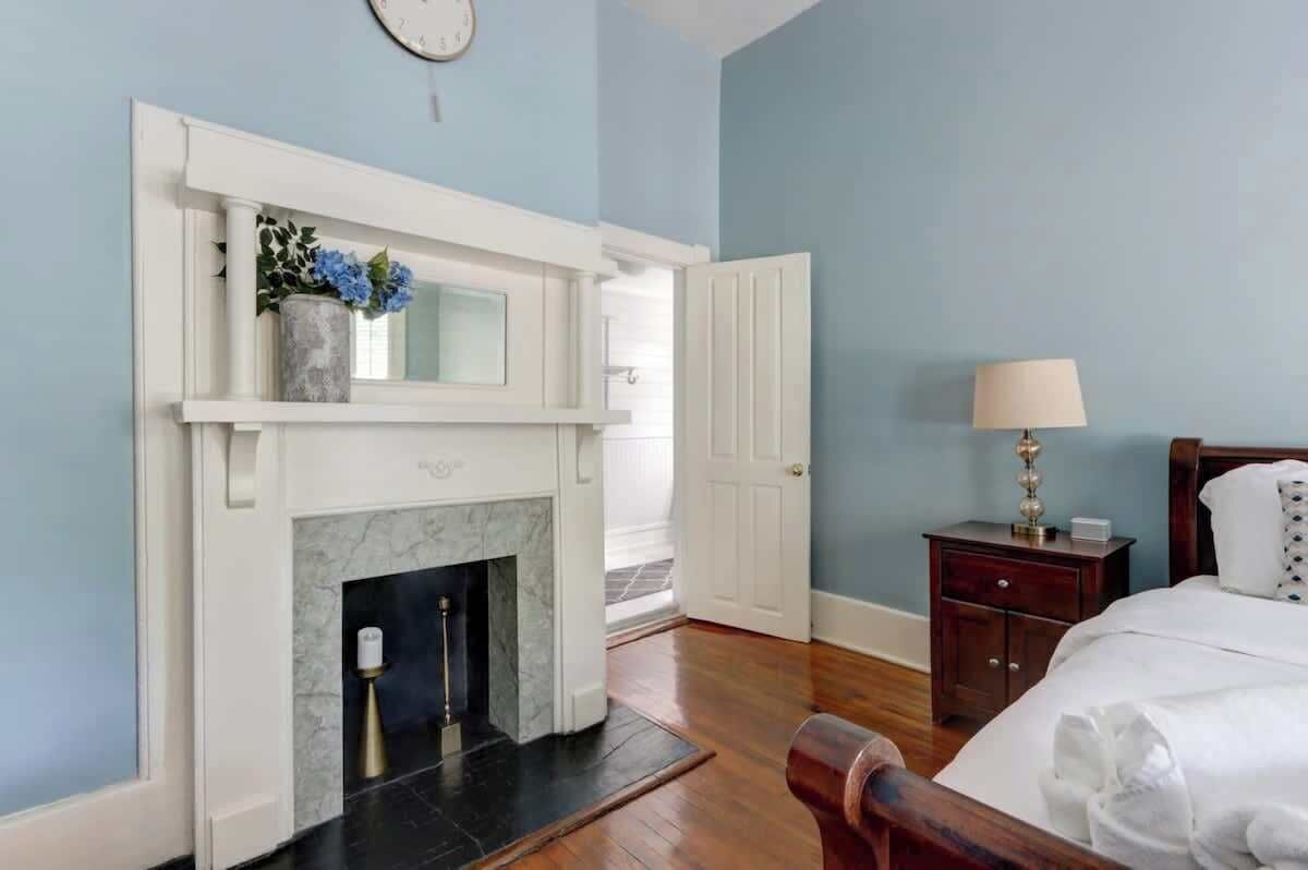 A bedroom with a white fireplace, a bed with white bedding, a nightstand with a lamp, and a wooden headboard.