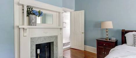 A bedroom with a white fireplace, a bed with white bedding, a nightstand with a lamp, and a wooden headboard.