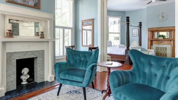 Une pièce avec une cheminée, deux fauteuils turquoise, une table ronde et un lit à l’arrière-plan.