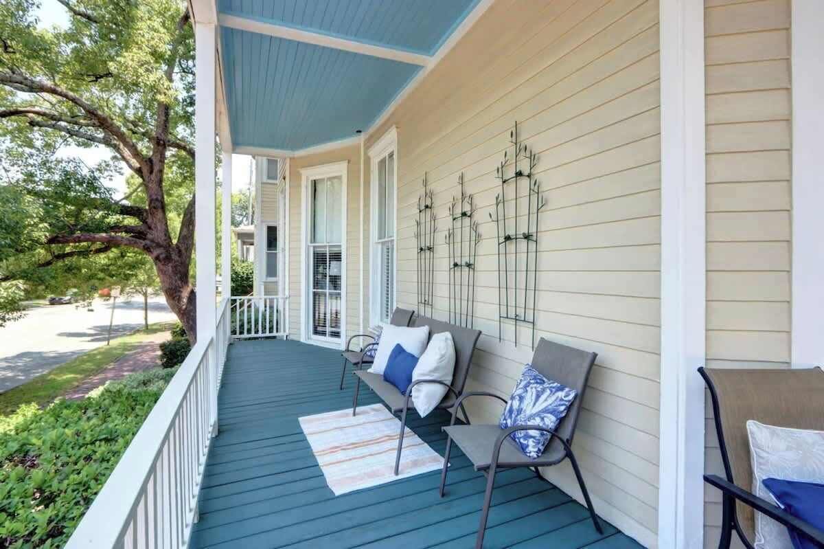A covered porch with a blue floor, white railings, and a row of chairs with cushions.