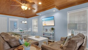 Een woonkamer met een plafondventilator, een eethoek met witte stoelen, een bruine leren bank en een schilderij van een strand.