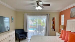A bedroom with a bed, a TV, a chair, a ceiling fan, and a view of an outdoor area.