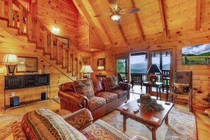 Interior de una cabaña de madera con un sofá de cuero, una mesa de comedor y una escalera que conduce a un balcón.