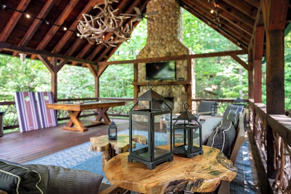 Une terrasse extérieure en bois, agrémentée d’une cheminée en pierre, d’une table ronde en bois et de deux lanternes.