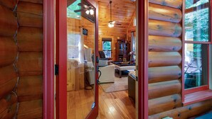 A log cabin interior with a wooden floor, a ceiling fan, a dining area with a table and chairs, and a view of the outdoors through a window. - Bentley's Retreat On Overlook (Blue Ridge)