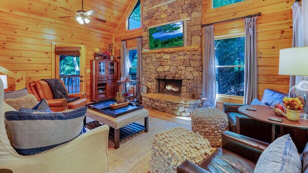 A cozy log cabin interior with a stone fireplace, leather furniture, and large windows offering views of the forest. - Bentley's Retreat On Overlook (Blue Ridge)