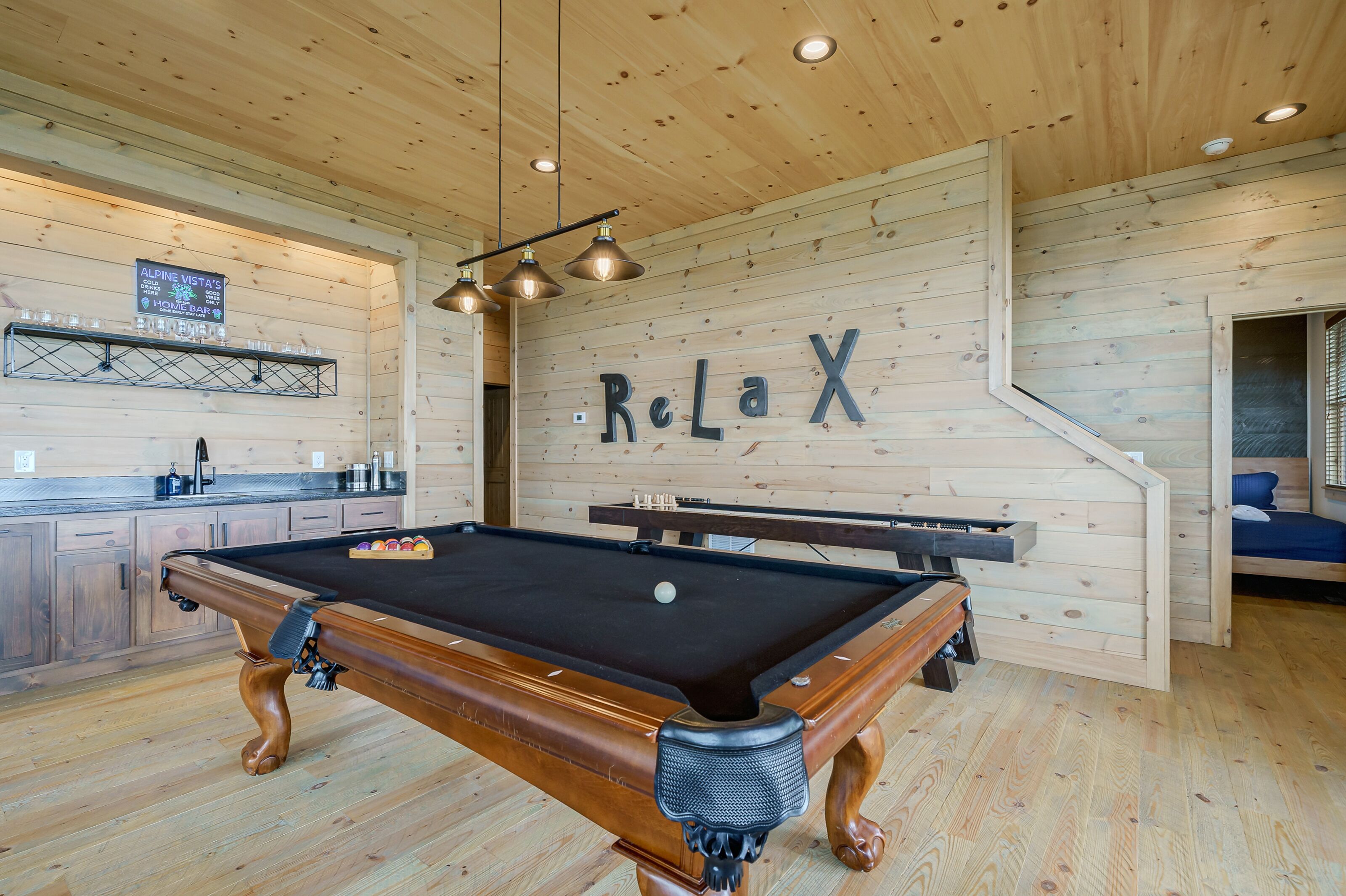 Une table de billard dans une pièce en bois, avec un panneau « Détendez-vous » accroché au mur et un coin bar où des verres sont posés sur une étagère.