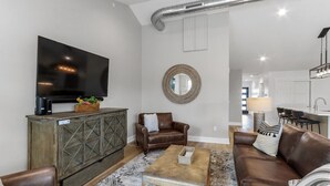 A modern living room with a large flat-screen TV, leather sofas, a coffee table, and a decorative mirror. - The Lofts of Brick and Mortar-City Loft |Stylish |Walk-Out Balcony |2 Kings |Parking for 2 |Downtown (Lake Geneva)