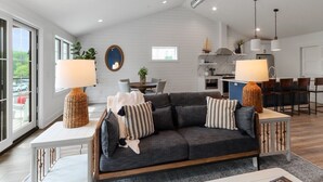 A modern living room with a grey sofa, two side tables with lamps, a dining area, and a kitchen in the background. - The Lofts of Brick and Mortar-Lake Loft |Luxury |Walk-Out Balcony |2 Kings |Parking for 2 |Downtown (Lake Geneva)