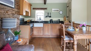 A kitchen with wooden cabinets, a dining table, and a stone wall. - Granary Cottage -  a cottage that sleeps 2 guests  in 1 bedroom (Kingsbridge)