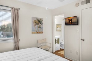 A bedroom with a bed, a chair, a painting, a television, and a bathroom visible through an open door. - Southbay 81 • Townhome w/ Seasonal Beach Service (Destin)