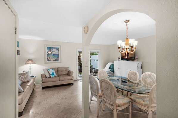 A dining area with a glass table and wicker chairs, a beige sofa, and a chandelier. - Southbay 81 • Townhome w/ Seasonal Beach Service (Destin)