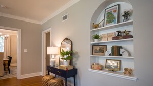A hallway with a dark blue console table, a mirror, and two woven ottomans. Shelves above contain decorative items and a framed map of Georgia.