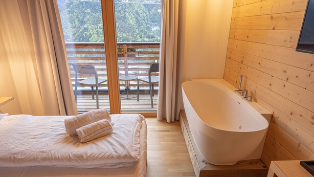 A bedroom with a large bed, a bathtub, and a view of a wooden deck and chairs outside. - B&B GIALLO - Felce Wellness Room (Pieve di Cadore)