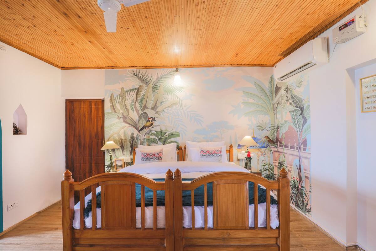 A bedroom with a wooden ceiling, a bed with white bedding, and a mural of tropical plants on the wall.